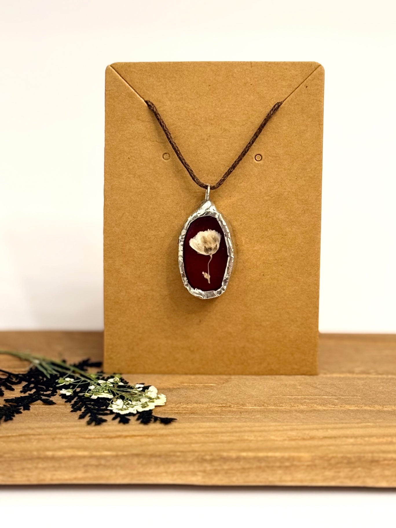 A dried flower pendant necklace with a silver foil frame against a dark red burgundy stained glass background with a dark brown cord, displayed on a brown kraft card stand on a wooden surface.