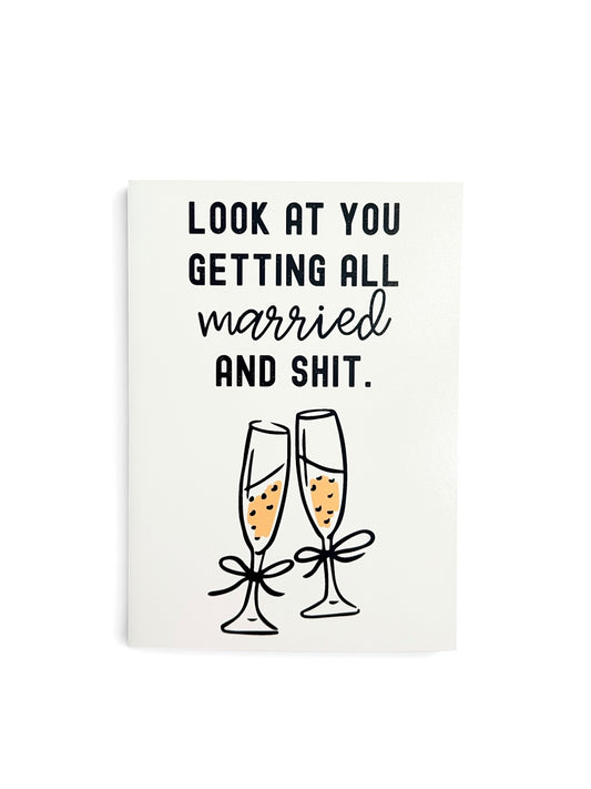 A white greeting card with black text reading 'LOOK AT YOU GETTING ALL married AND SHIT.' above an illustration of two champagne flutes with yellow champagne and black bows.