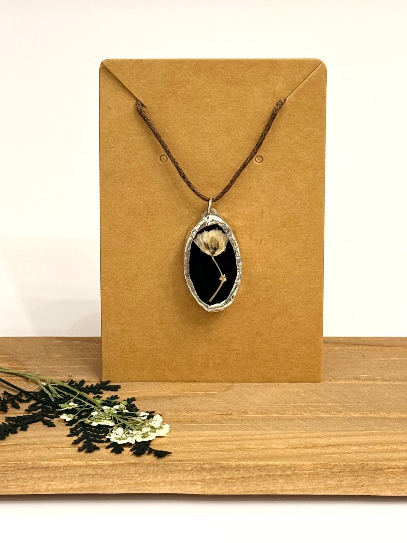 A dried flower black stained glass pendant necklace displayed on a brown kraft card, set on a wooden surface with dried flowers in the foreground.