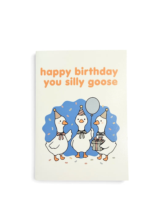 A vertical greeting card featuring three cartoon white geese wearing party hats and scarves, with one goose holding a silver balloon and another holding a gift box. The card has orange text at the top reading 'happy birthday you silly goose' on a white background, set against a clean white studio backdrop.