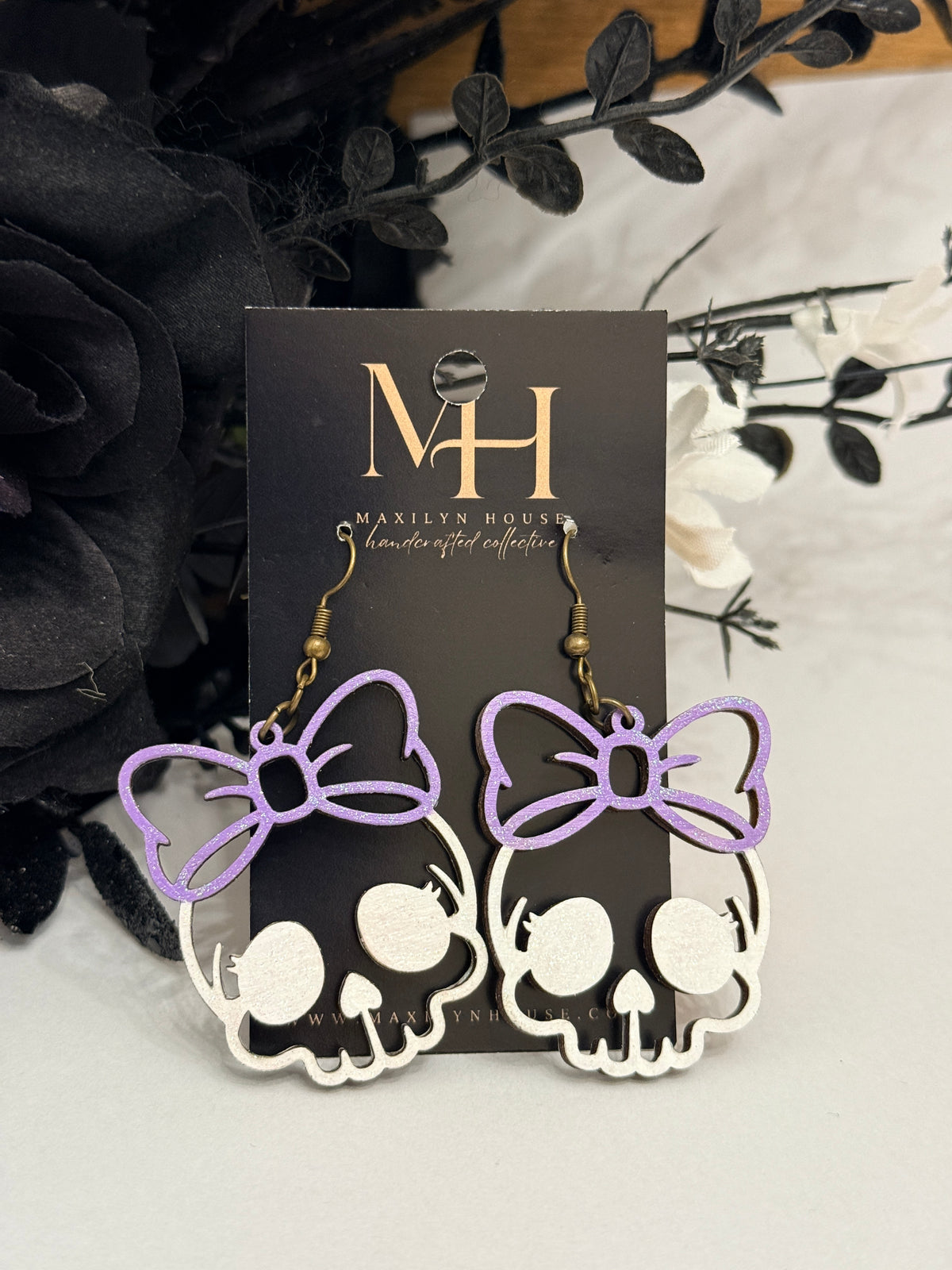 A pair of dangling earrings displayed on a black branded card. Each earring features a white skull silhouette with a lavender purple bow outline above it and gold-tone hooks.