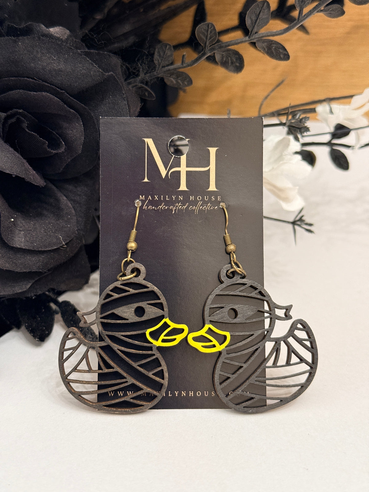 A pair of duck shaped drop earrings with a black body and bright yellow bill. Displayed on a black card labeled MH. The earrings are styled with black and white artificial flowers on a light surface.