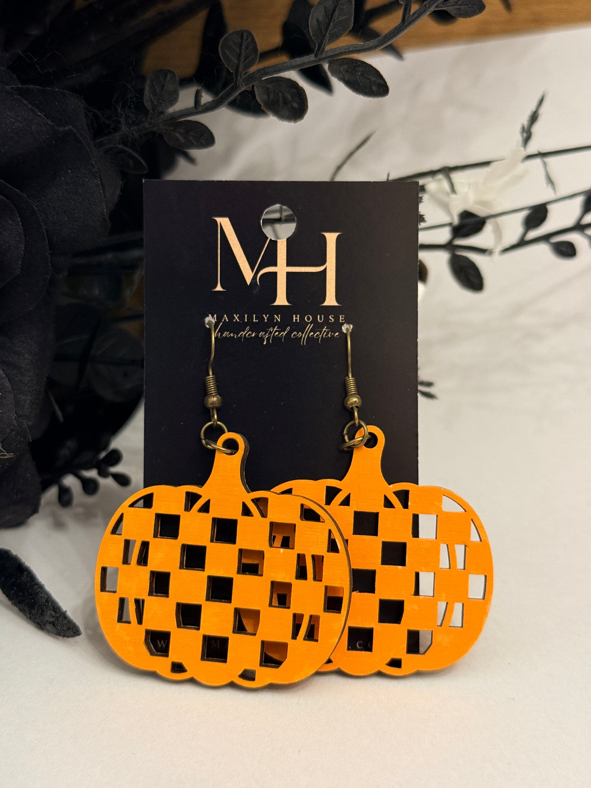 Bright orange pumpkin-shaped dangle earrings with a checkerboard cutout pattern, displayed on a black card labeled MH. The earrings are styled with dark decorative leaves on a light surface.