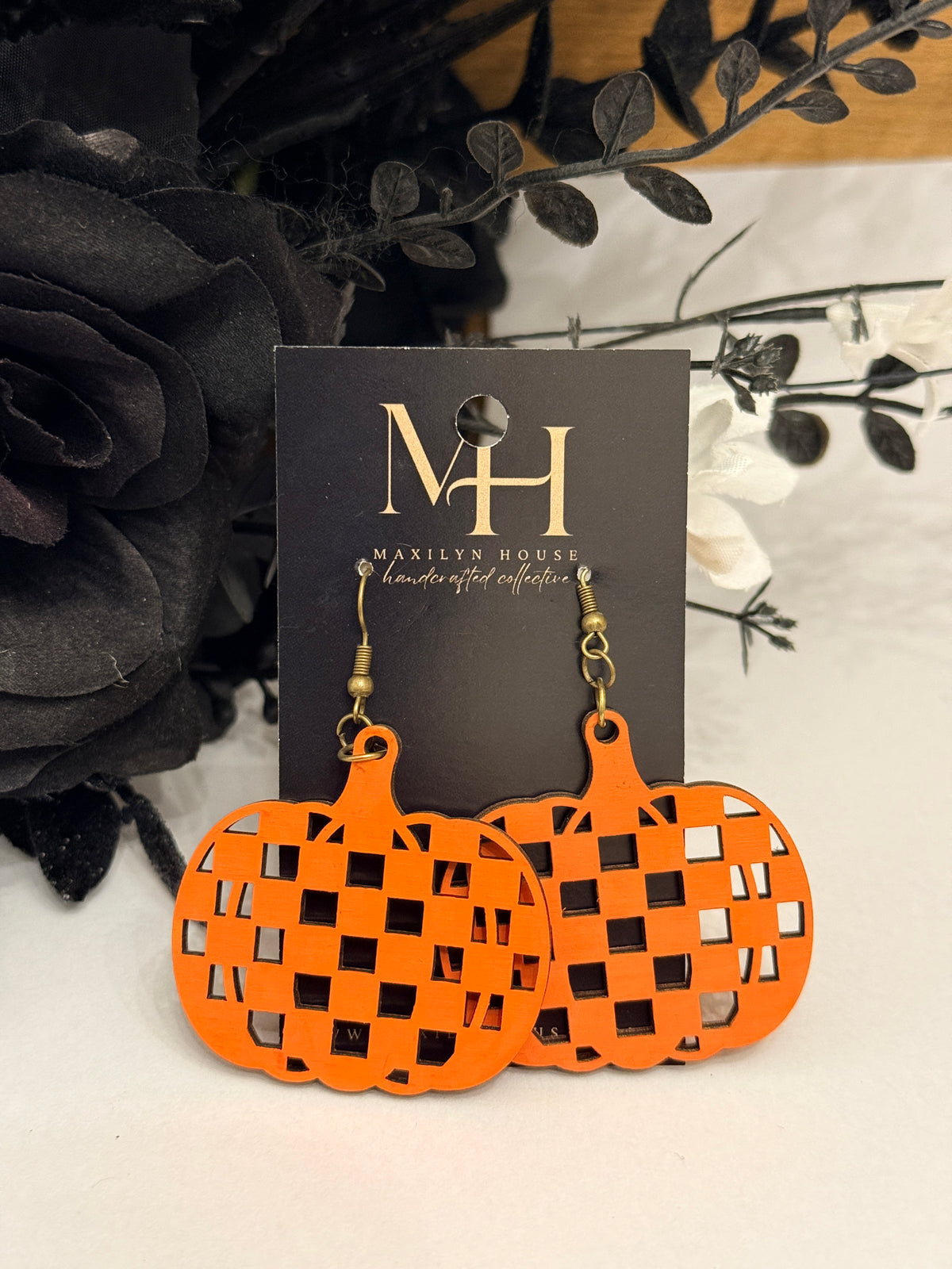 Orange pumpkin-shaped dangle earrings with a checkered cutout pattern, displayed on a black card labeled MKELTY HOUSE, with black and white decorative flowers in the background.