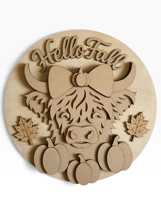 A circular wood and mdf laser-cut decorative sign featuring a Highland cow with a bow, the words 'Hello Fall' in script, and several pumpkins, shown on a plain white background.