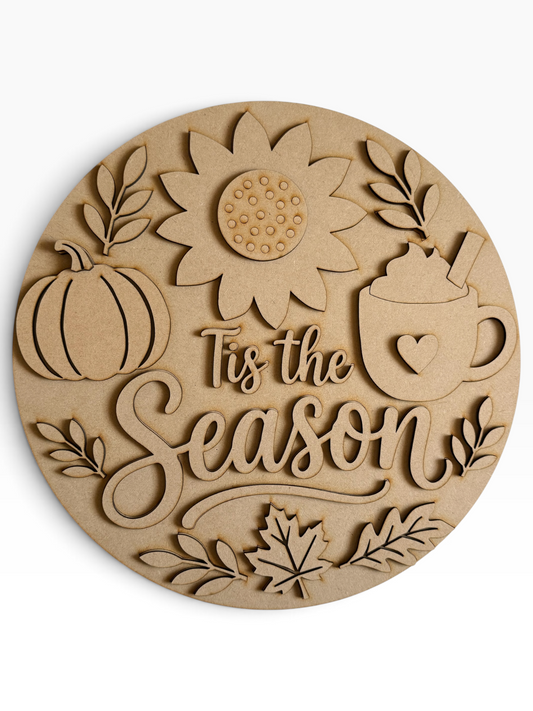 A round laser-cut mdf round sign featuring autumn-themed designs including a pumpkin, sunflower, leaves, a hot cocoa mug with a heart, and the phrase 'Tis the Season' in elegant script, shown on a clean white background.