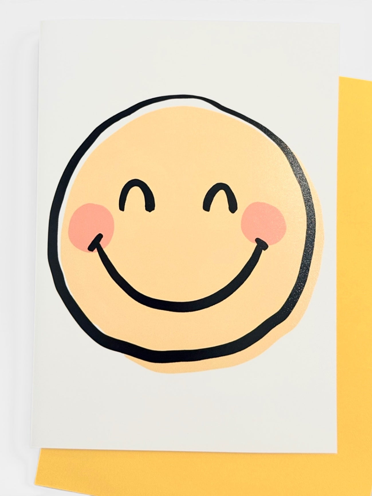 A vertical white greeting card featuring a minimalist smiley face with a yellow face, black outline, and pink cheeks, layered over a yellow envelope.