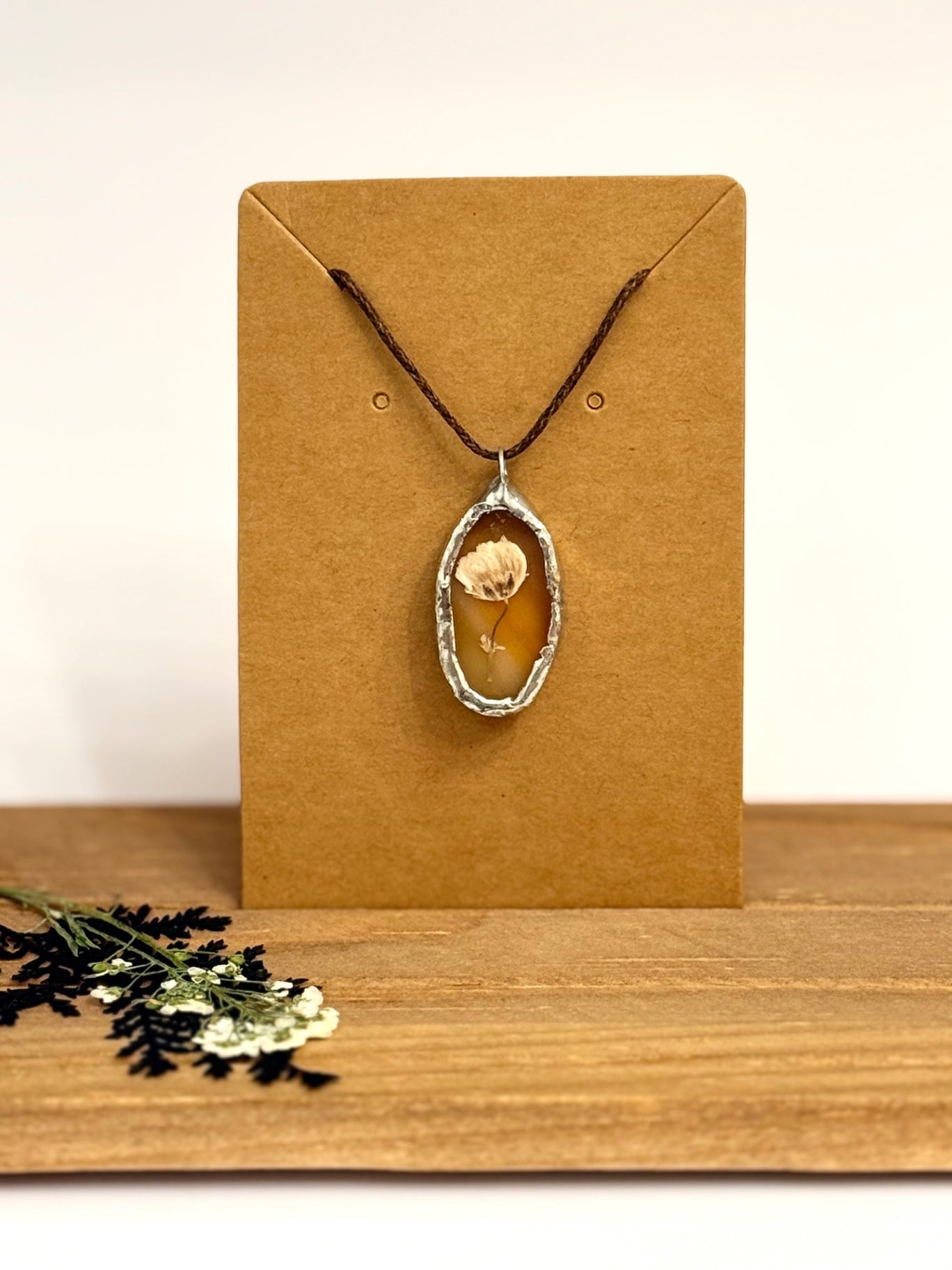 A dried flower pendant necklace with a silver toned foil setting against an orange stained glass background, with dark cord, displayed on a brown kraft card  with a wooden surface and small dried flowers in the foreground.