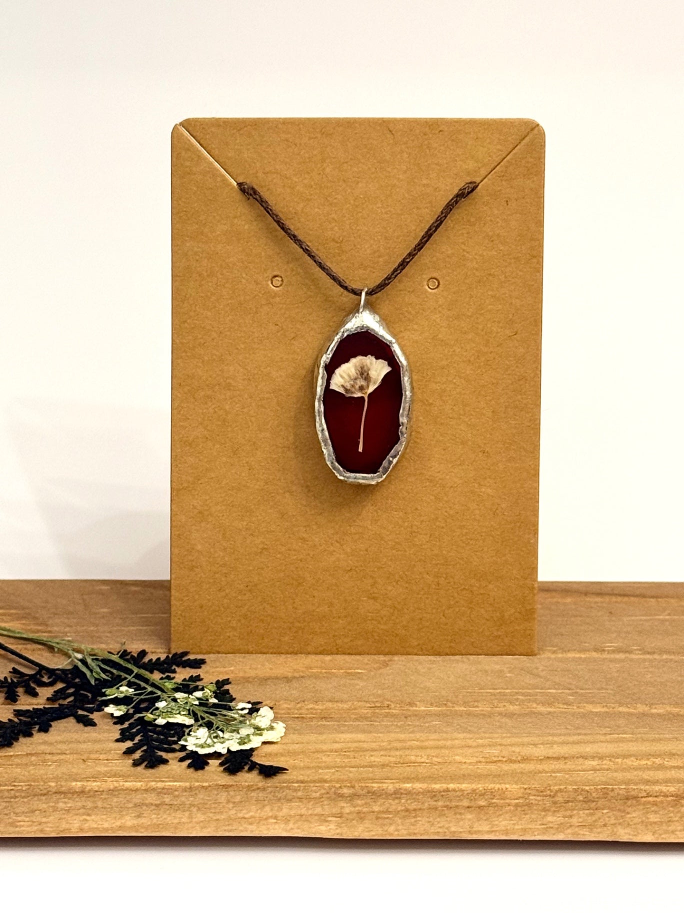 A dried flower pendant necklace with a burgundy stained glass center and silver foil border, displayed on a brown kraft card. The necklace is hung on a dark brown cord, set against a neutral background with dried flowers on a wooden surface.