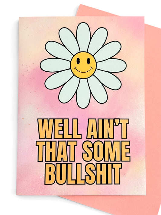 A greeting card with a pink watercolor background, a smiling daisy flower with a yellow face, and bold yellow text reading 'WELL AIN'T THAT SOME BULLSHIT'. A pink envelope is partially visible behind the card.