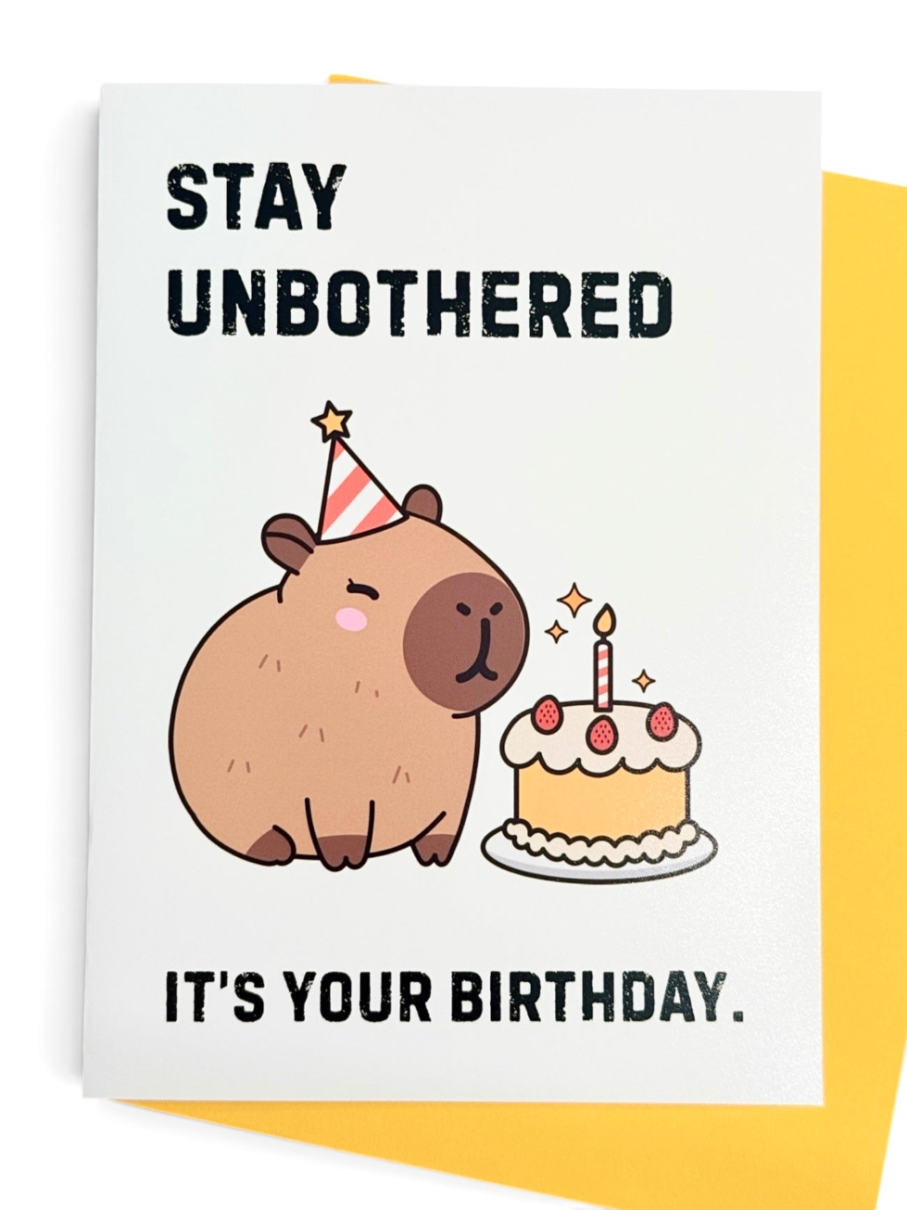 A rectangular card with a white background featuring a cute cartoon capybara wearing a party hat next to a birthday cake, with the text 'STAY UNBOTHERED' at the top and 'IT'S YOUR BIRTHDAY.' at the bottom. Yellow card envelope shown behind the card. 
