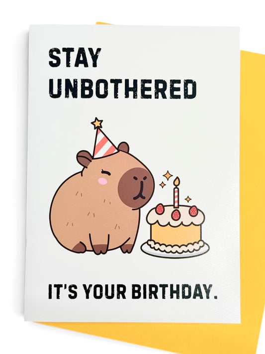 A rectangular card with a white background featuring a cute cartoon capybara wearing a party hat next to a birthday cake, with the text 'STAY UNBOTHERED' at the top and 'IT'S YOUR BIRTHDAY.' at the bottom. Yellow card envelope shown behind the card. 