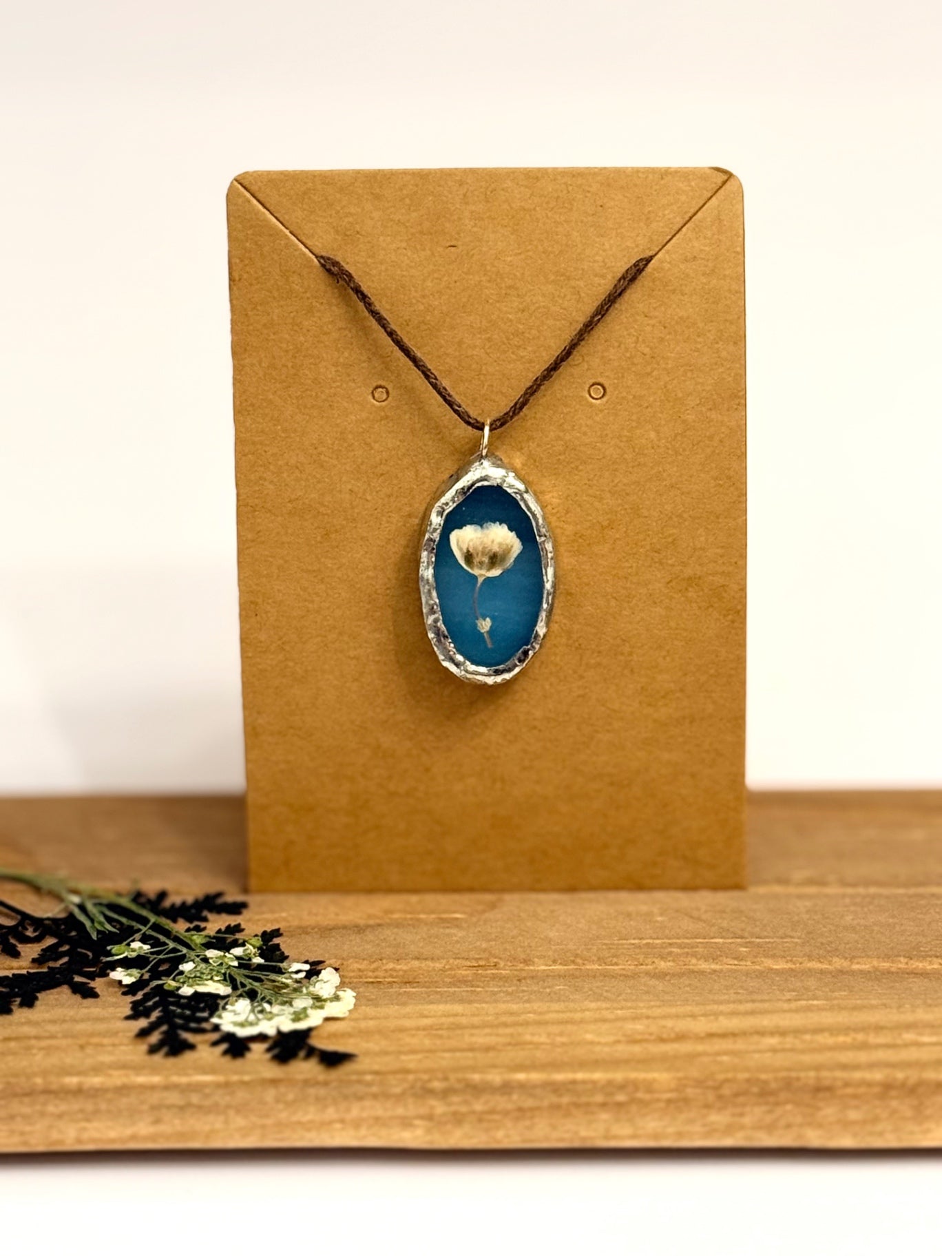 A silver foil set oval pendant necklace with a blue stained glass background and a dried flower inside, displayed on a brown kraft card stand on a wooden surface.