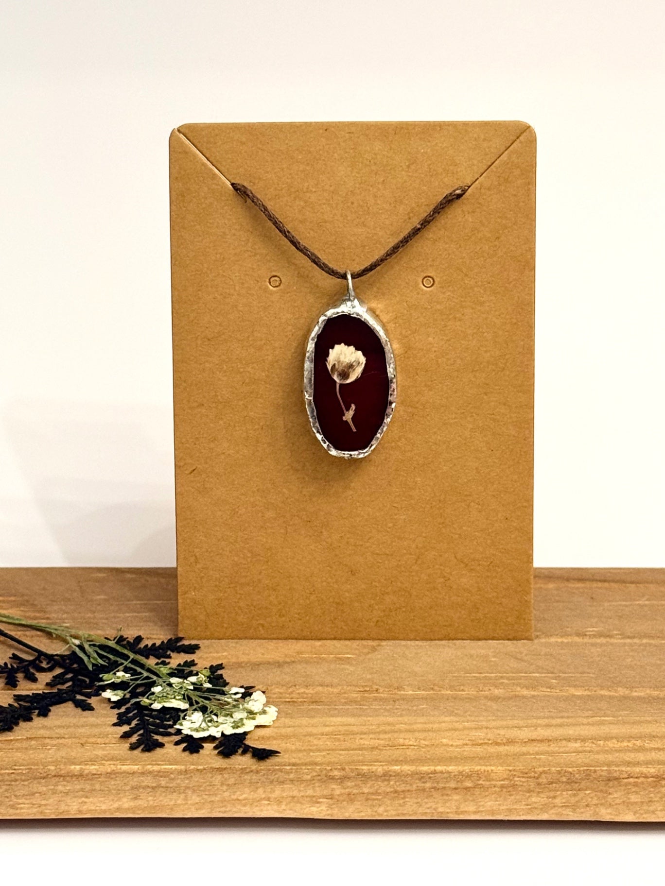 A dried flower pendant necklace set in foil with a burgundy stained glass background with brown cord, displayed on a brown kraft card, set on a wooden surface with dried flowers in the foreground.