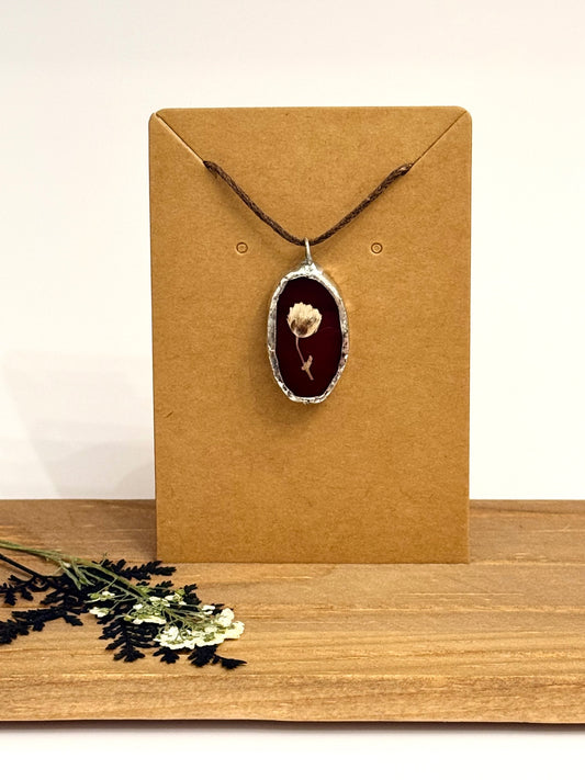 A dried flower pendant necklace set in foil with a burgundy stained glass background with brown cord, displayed on a brown kraft card, set on a wooden surface with dried flowers in the foreground.