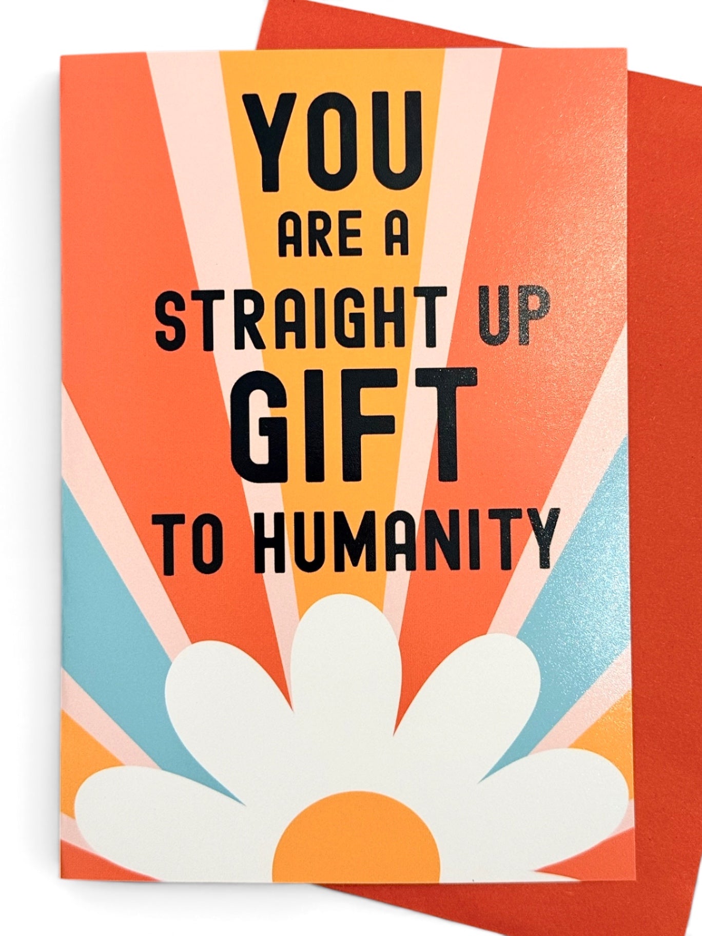 A rectangular vertical  card with retro-style text reading 'YOU ARE A STRAIGHT UP GIFT TO HUMANITY' in black over orange, peach, cream, and light blue rays, with a large white daisy flower at the bottom, shown on a white background. A matching orange envelope is partially visible behind the card.