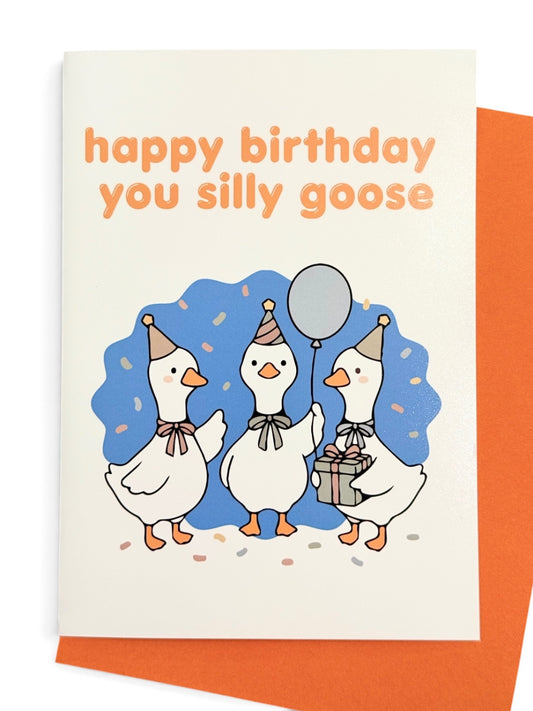 A vertical greeting card with an orange envelope, featuring three cartoon white geese wearing party hats and scarves, with one goose holding a silver balloon and another holding a gift box. The card has orange text at the top reading 'happy birthday you silly goose' on a white background, set against a clean white studio backdrop.