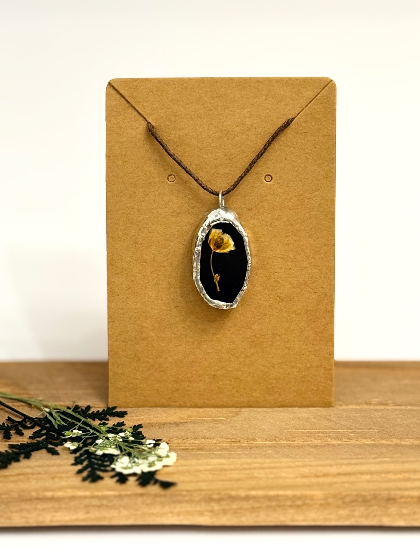 A silver foil pendant necklace featuring a dried yellow flower on a black stained glass background, displayed on a brown kraft card. The card is set on a wooden surface with dried greenery in the foreground.