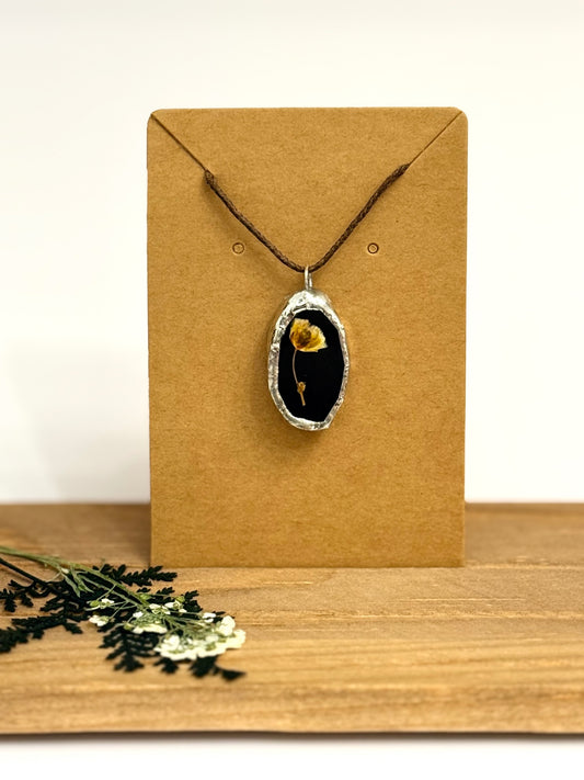 A silver foil pendant necklace featuring a dried yellow flower on a black stained glass background, displayed on a brown kraft card. The card is set on a wooden surface with dried greenery in the foreground.