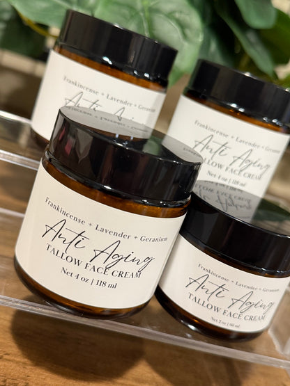 Four amber glass jars of Anti Aging Tallow Face Cream with black lids and white labels, arranged on a clear acrylic stand over a wooden surface with green leaves in the background.