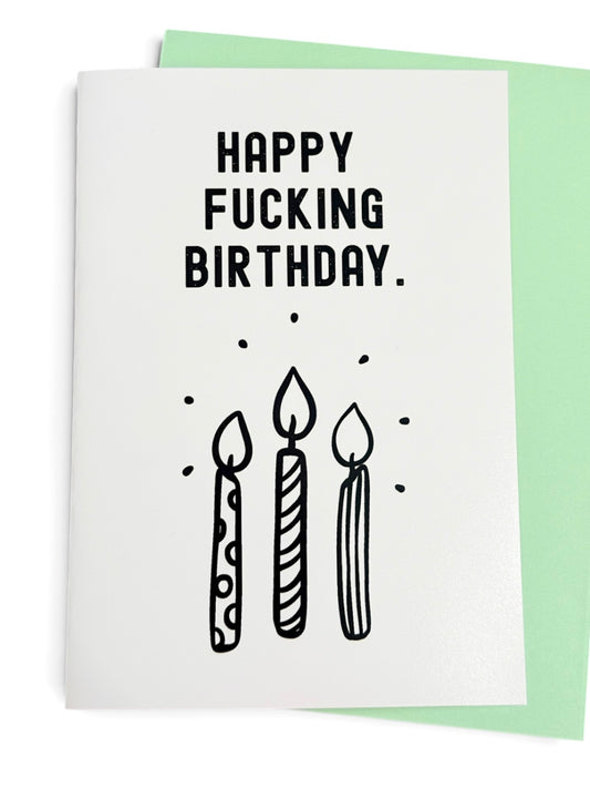 A white greeting card with the text 'HAPPY FUCKING BIRTHDAY.' in bold black letters above a simple black line drawing of three candles, shown with a light green envelope behind it on a white background.