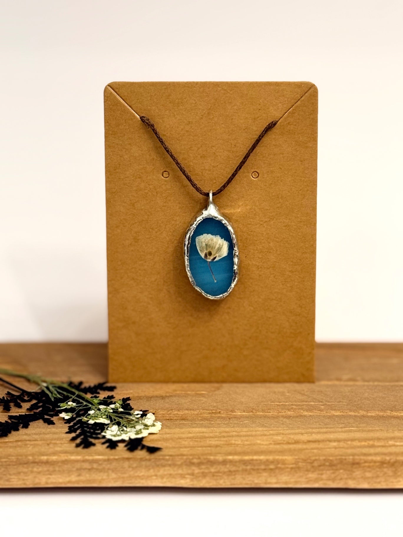A silver foil set oval pendant necklace with a blue stained glass background and a small white flower in the center, hanging from a dark cord on a brown kraft display card. The card is set on a wooden surface against a plain light background, with dried flowers in the lower left corner.