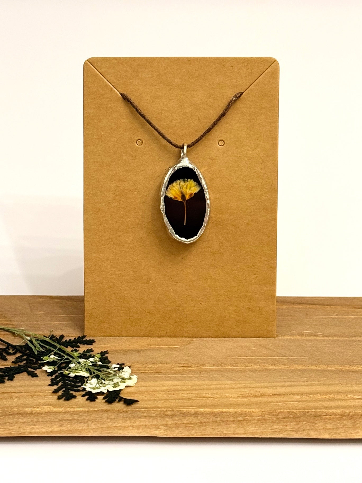 A silver oval pendant necklace featuring a dried yellow flower on a black stained glass background, displayed on a brown kraft card. The card is set on a wooden surface with dried flowers in the foreground.