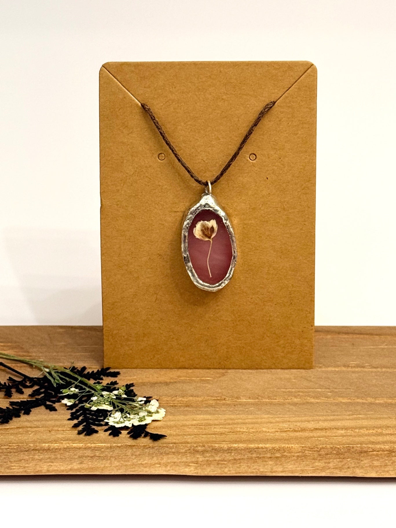A dried flower pendant necklace with a silver-foil rimmed oval frame against mauve colored stained glass and dark brown cord, displayed on a brown kraft card stand on a wooden surface.