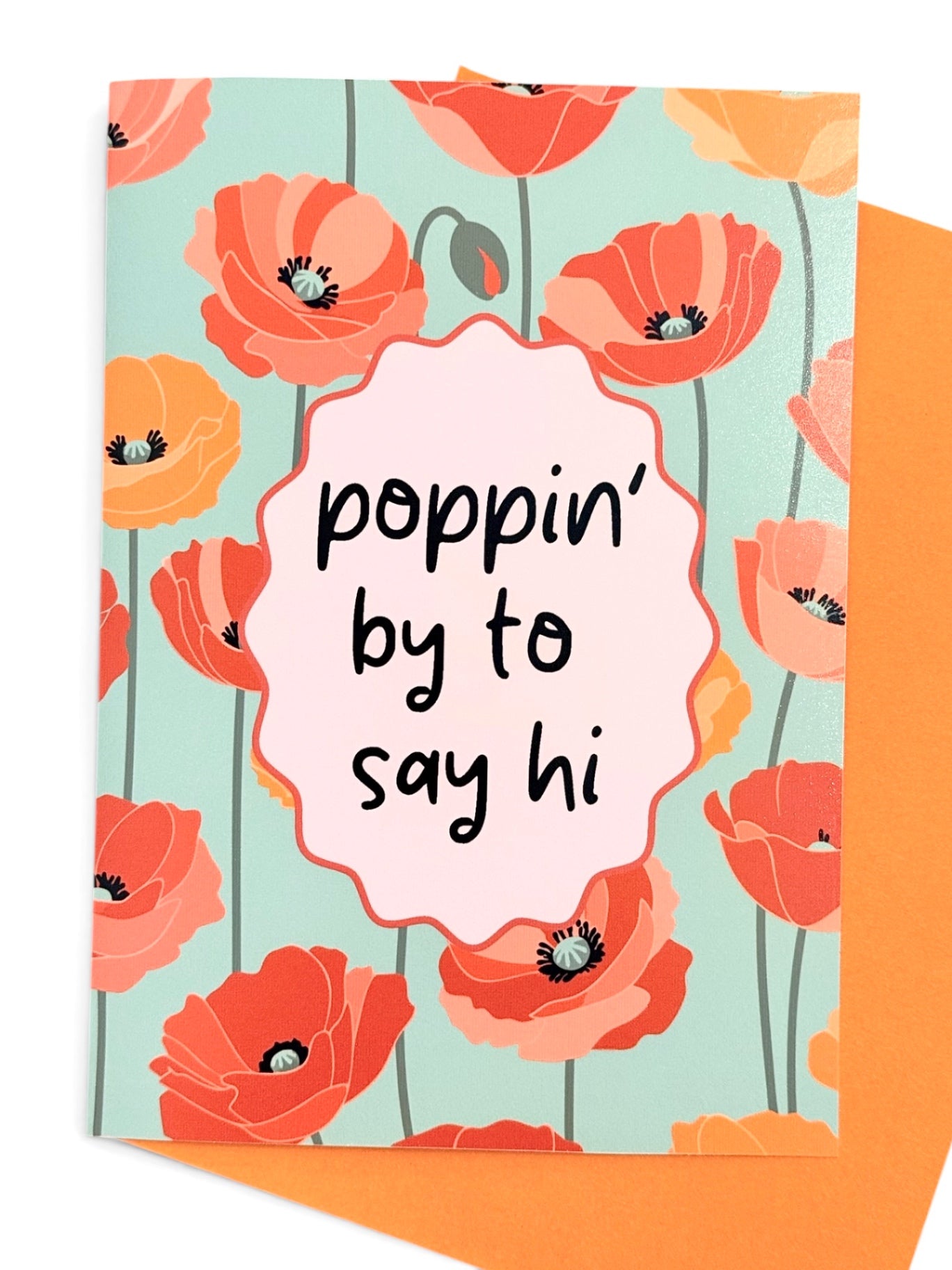A greeting card with a floral poppy pattern in coral, orange, and peach on a light aqua background, featuring a central pink scalloped label with the handwritten text 'poppin' by to say hi'. An orange envelope is partially visible behind the card.