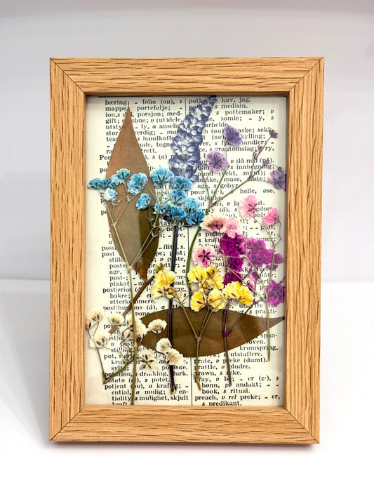 A rectangular wooden frame containing pressed dried flowers and leaves arranged over a vintage book page background, photographed against a light gray studio backdrop.