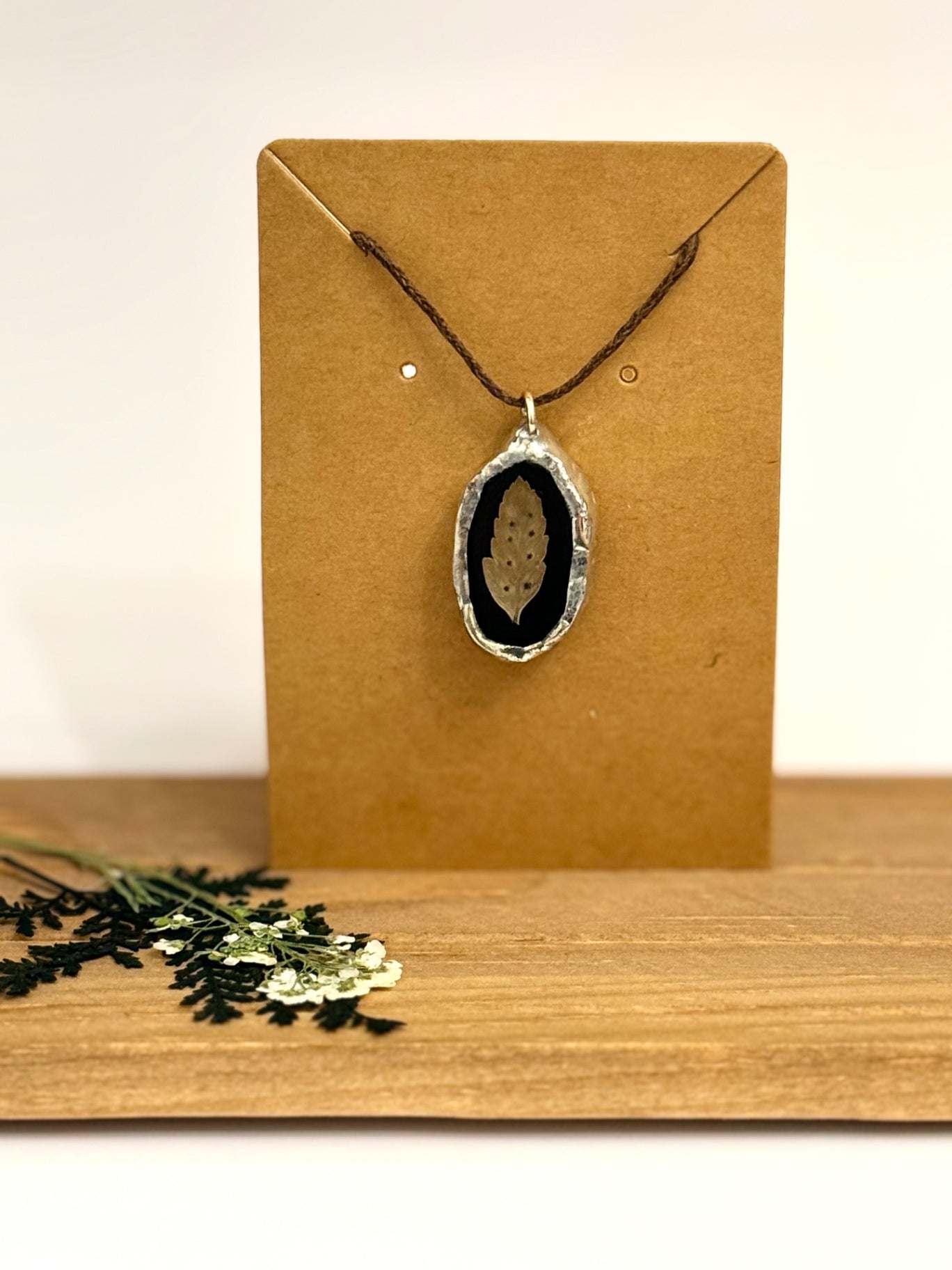 A silver foil pendant necklace with a dried floral leaf set against a black stained glass background, displayed on a brown kraft card stand on a wooden surface.
