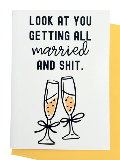 A white greeting card with black text reading 'LOOK AT YOU GETTING ALL married AND SHIT.' above an illustration of two champagne flutes with yellow champagne and black bows, shown with a yellow envelope behind it.