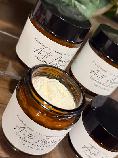 Several amber glass jars of tallow face cream with black lids and white labels are arranged on a wooden surface. One jar in the foreground is open, showing a pale yellow cream inside, while the others are closed.