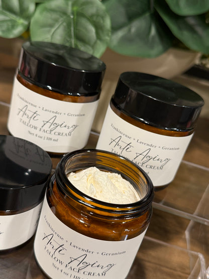 Several amber glass jars of face cream with black lids and white labels reading 'Anti Aging Tallow Face Cream' are arranged on a tray, with one jar open to show the cream inside. Green leaves are visible in the background.