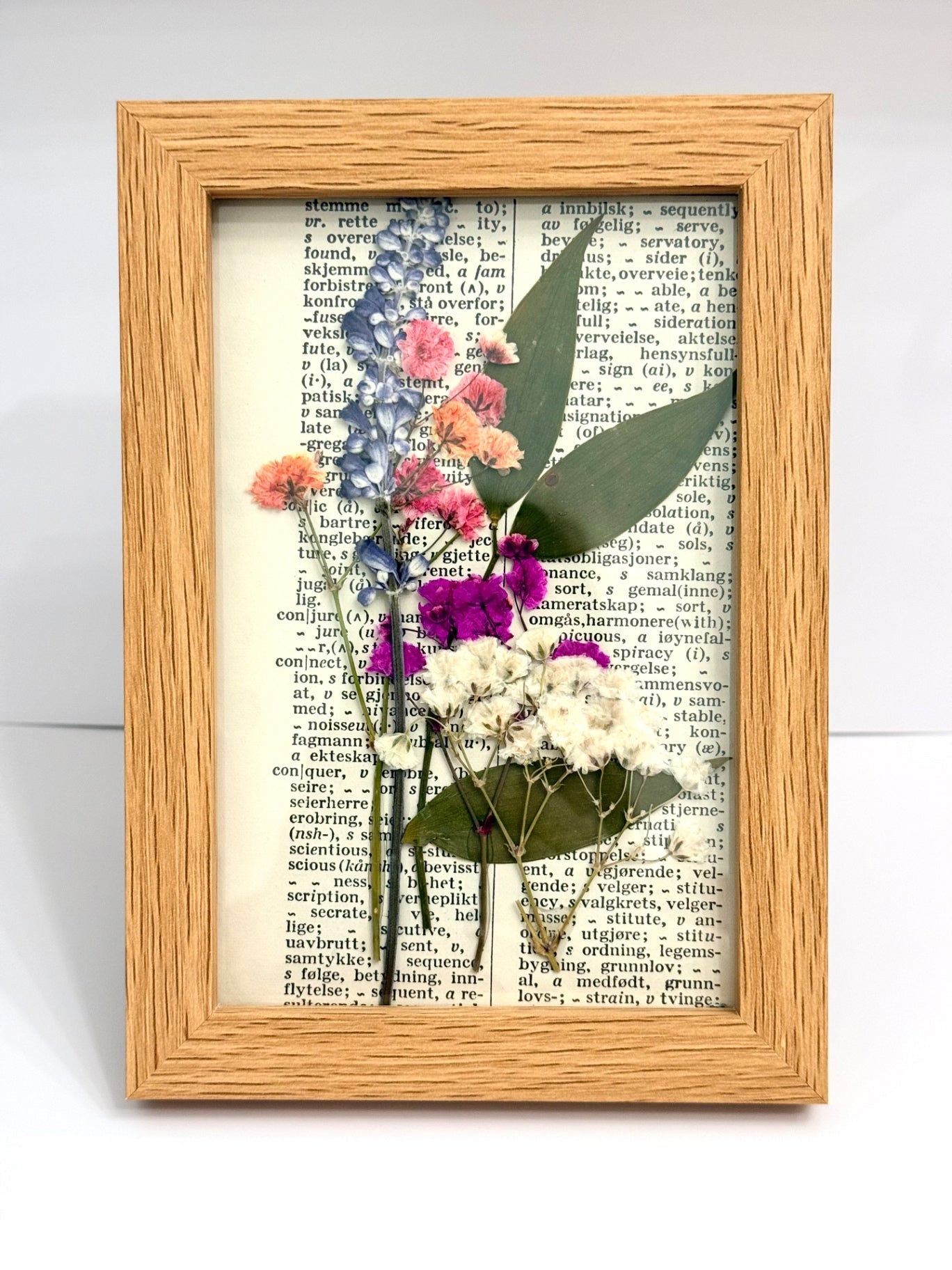 A framed botanical art piece featuring pressed flowers in shades of purple, pink, and white with green leaves, arranged over a vintage dictionary page background inside a light wood frame.