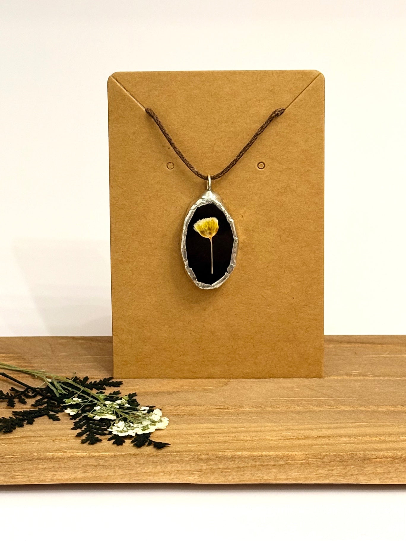 A pendant necklace with a dried yellow flower set in a black stained glass oval and silver foil bezel, displayed on a brown kraft card stand on a wooden surface.