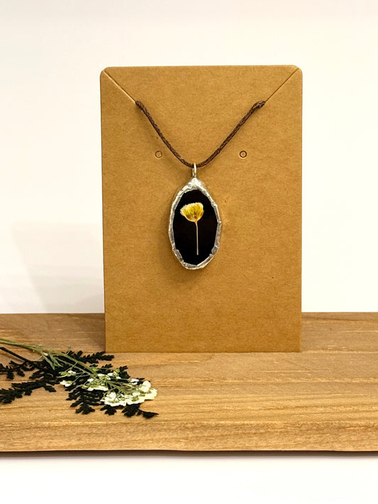 A pendant necklace with a dried yellow flower set in a black stained glass oval and silver foil bezel, displayed on a brown kraft card stand on a wooden surface.