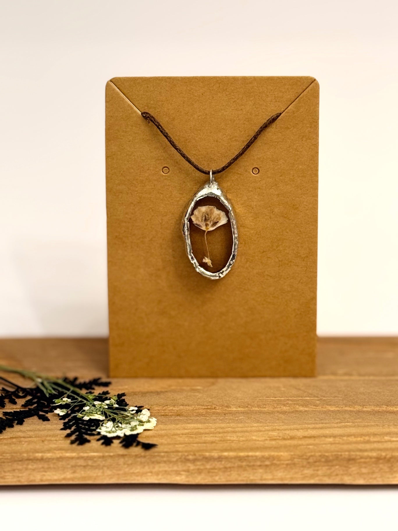 A silver foil setted pendant necklace with a dried flower preserved inside set against clear stained glass, hanging from a dark cord on a brown kraft display card. The card is set on a wooden surface with dried flowers in the foreground against a plain light background.