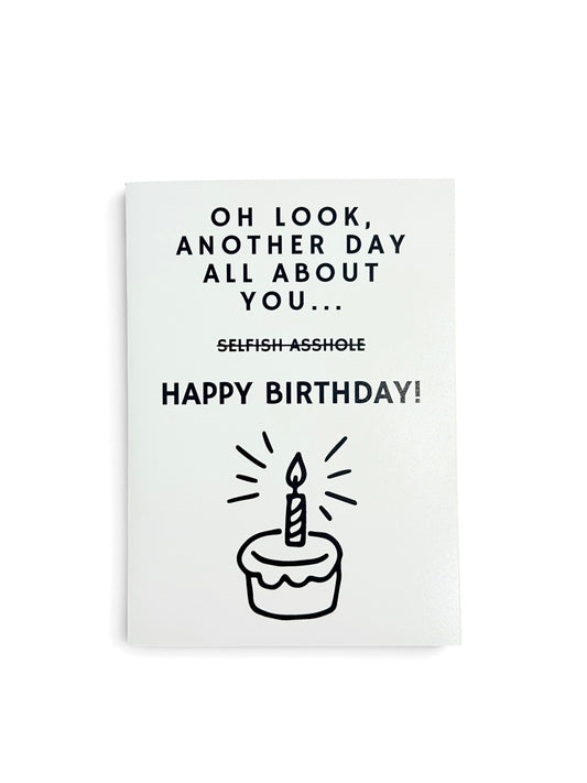 A white greeting card with black text that reads 'OH LOOK, ANOTHER DAY ALL ABOUT YOU with the words SELFISH ASSHOLE crossed out then a HAPPY BIRTHDAY greeting and an illustration of a cake with one candle burning. 