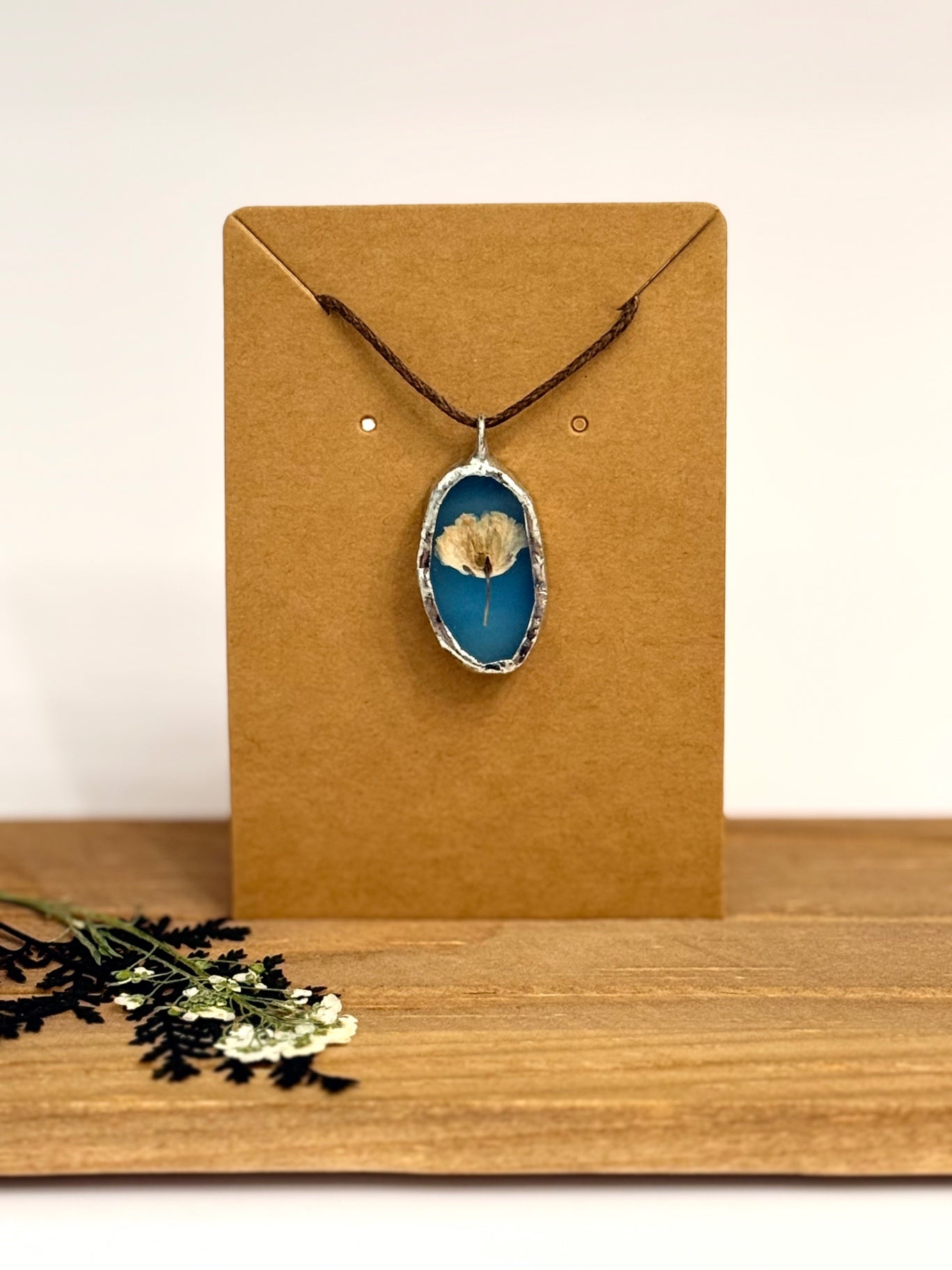 A foil set oval pendant necklace with a blue stained glass background and a dried flower in the center, hanging from a dark cord on a brown kraft display card. The card is set on a wooden surface against a light neutral background.