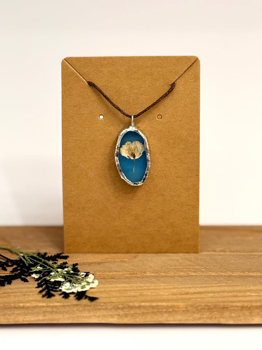 A foil set oval pendant necklace with a blue stained glass background and a dried flower in the center, hanging from a dark cord on a brown kraft display card. The card is set on a wooden surface against a light neutral background.