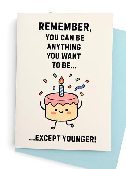 A white greeting card with a cute cartoon birthday cake character and the text 'REMEMBER, YOU CAN BE ANYTHING YOU WANT TO BE..' above the cake then text 'EXCEPT YOUNGER' below the cake. Shown with a light blue colored envelope behind the card.