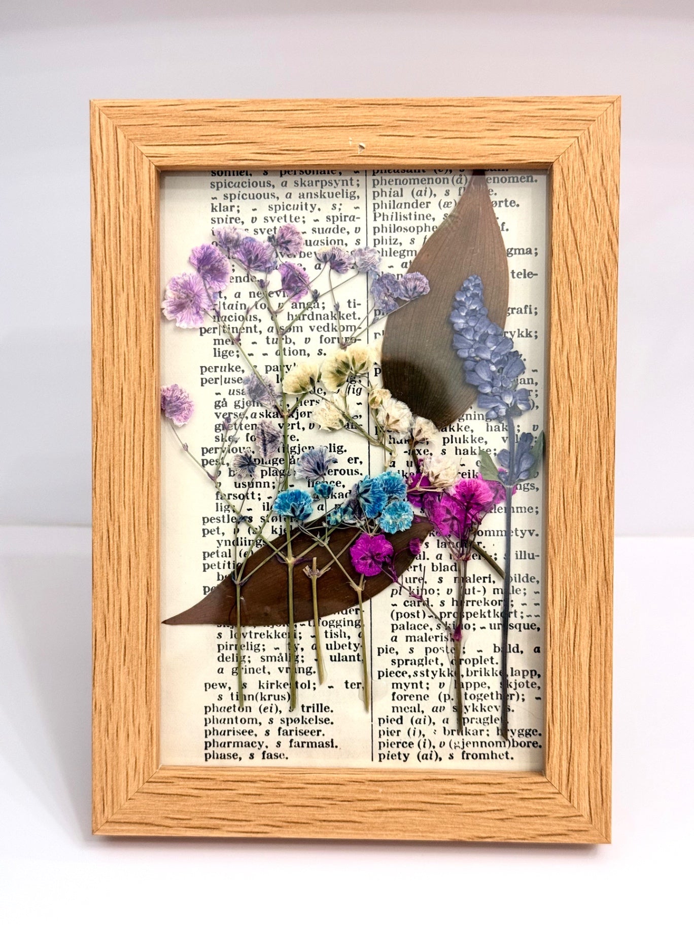 A rectangular wooden shadow box frame containing a vintage book page background, decorated with dried flowers in purple, blue, and white, and a brown silhouette of a woman, set against a plain white background.