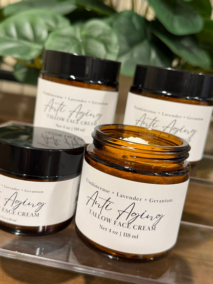 Four amber glass jars of Anti Aging Tallow Face Cream with black lids and white labels, arranged on a clear acrylic stand with green foliage in the background.