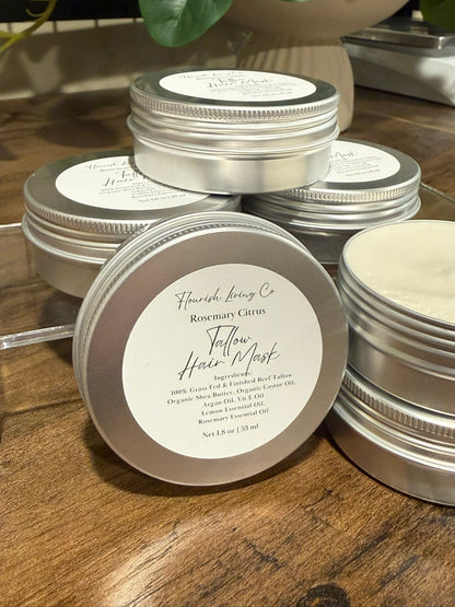 Several small round metal tins of hair masks are arranged on a wooden surface. The front tin is open, showing a pale cream-colored product inside, while the others are closed with white labels.