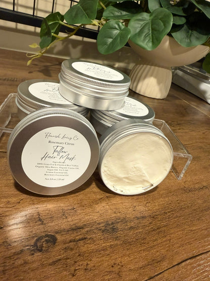Several round silver metal tins of hair product arranged on a wooden tabletop, with one tin open to show a pale cream-colored product inside. The front tin label reads 'FlourishLiving Co'