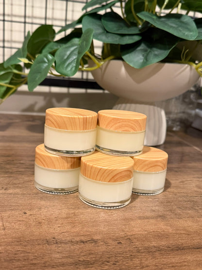Five small glass jars filled with off-white cream or balm, each topped with a light natural wood lid, arranged on a wooden surface with a potted green plant in the background.