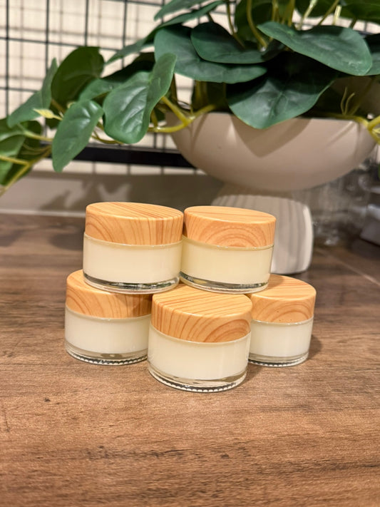 Five small glass jars filled with off-white cream or balm, each topped with a light natural wood lid, arranged on a wooden surface with a potted green plant in the background.