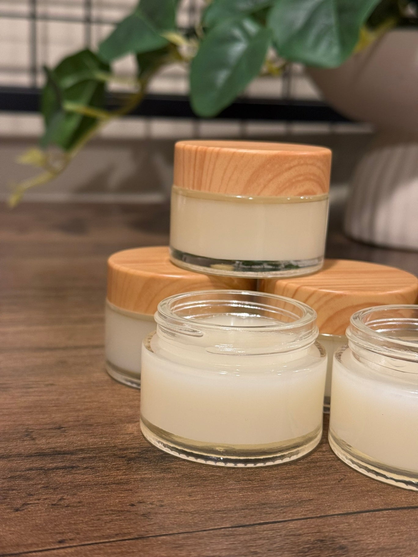 Several small clear cosmetic jars filled with a creamy off-white product, arranged on a wooden surface. Each jar has a light natural wood lid.