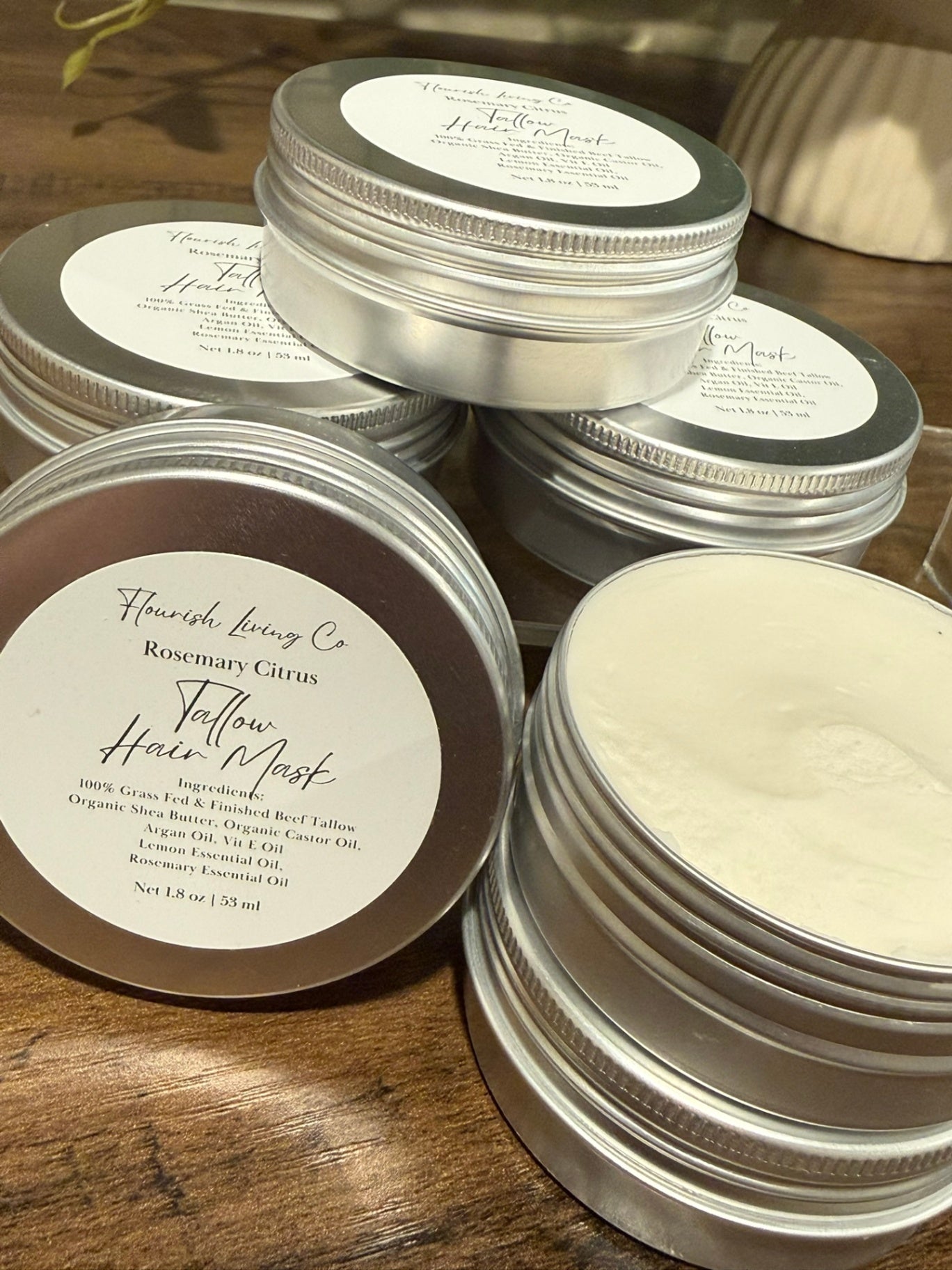 Several round silver metal tins of hair masks are arranged on a wooden surface. One tin is open, showing a white cream product, while others are closed with white labels.